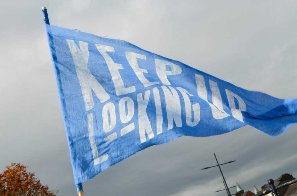A blue fabric flag waves in the wind with 'Keep Looking Up' painted across it. 