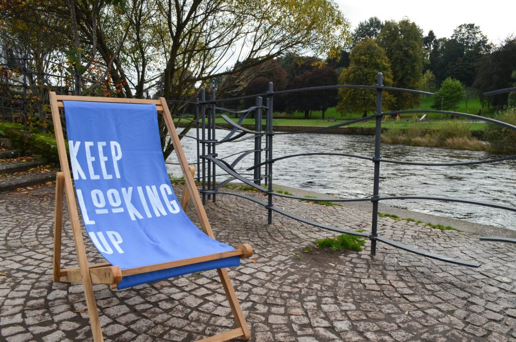 A blue deckchair with 'Keep Looking Up' painted on it, sitting on cobblestones next to a river. 