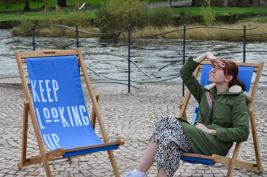 A person sitting on a blue deckchair next to the river, wearing a green jacket, looks to the sky. Another deckchair is next to them which has 'Keep Looking Up' painted across it. 