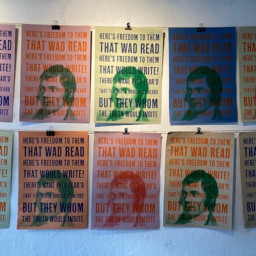 Wall display of printed posters arranged in a grid. Each poster repeats the same bold, capitalised text over a semi‑transparent, stylised portrait in varying colours, including green, red, and blue. The text reads: “THERE’S FREEDOM TO THEM THAT WAD READ / HERE’S FREEDOM TO THEM THAT WAD WRITE! / THERE’S NONE EVER FEAR’D THE TRUTH WOULD INDITE / BUT THEY WHOM.” The design uses contrasting colours and layered imagery to create a striking visual effect.