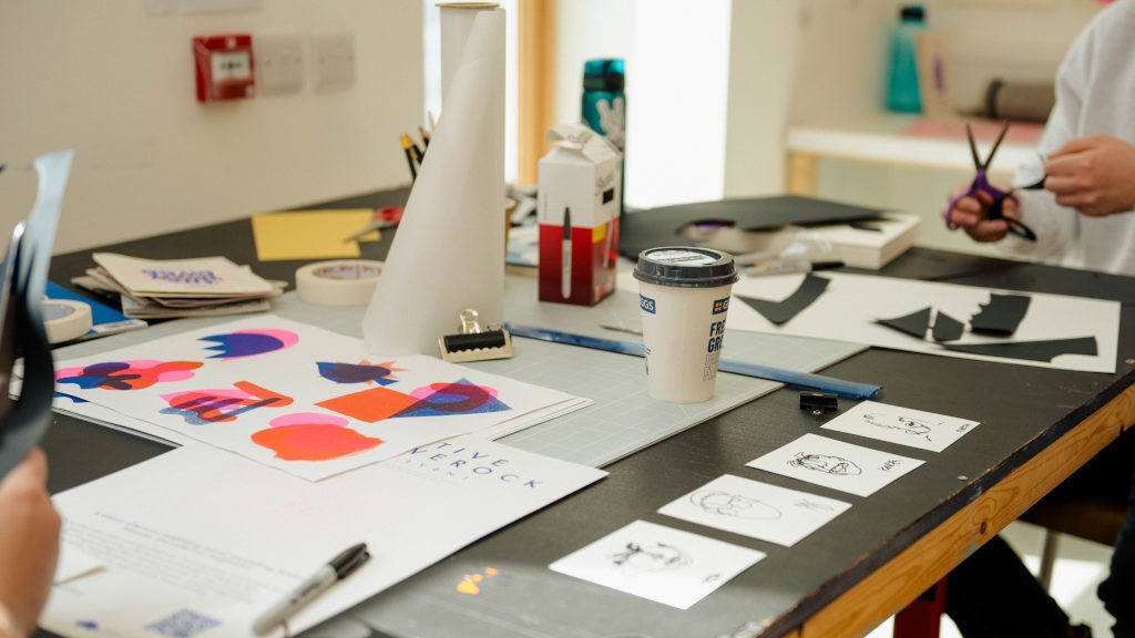 Two people working at a large table covered with art and craft materials, including colorful paper cutouts, black‑and‑white sketches, scissors, a coffee cup, and a cutting mat. Several small illustrations are arranged in a row, suggesting a creative design or illustration project in progress.