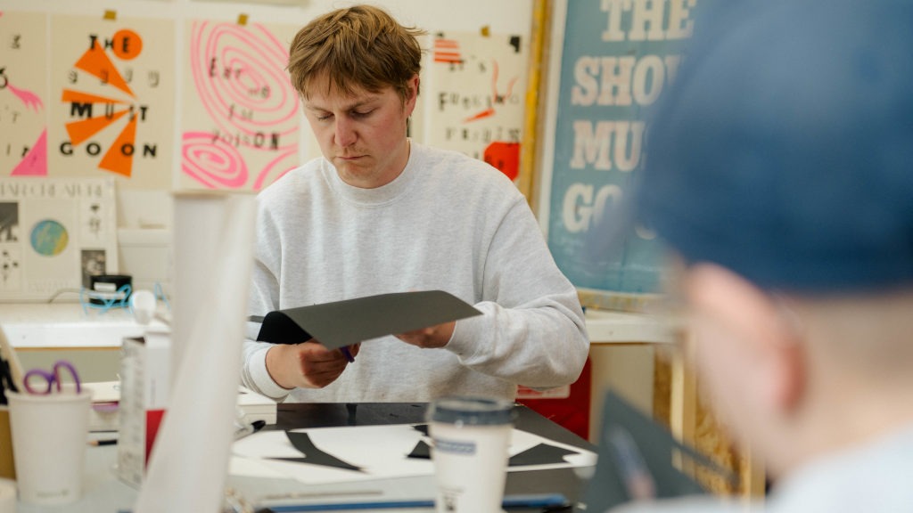 Person seated at a table in a creative studio, cutting a piece of black paper. The table is scattered with art supplies, including white paper with black shapes, scissors, and cups. Behind them, a wall displays posters and abstract artwork, including one with large text reading “THE SHOW MUST GO ON.”