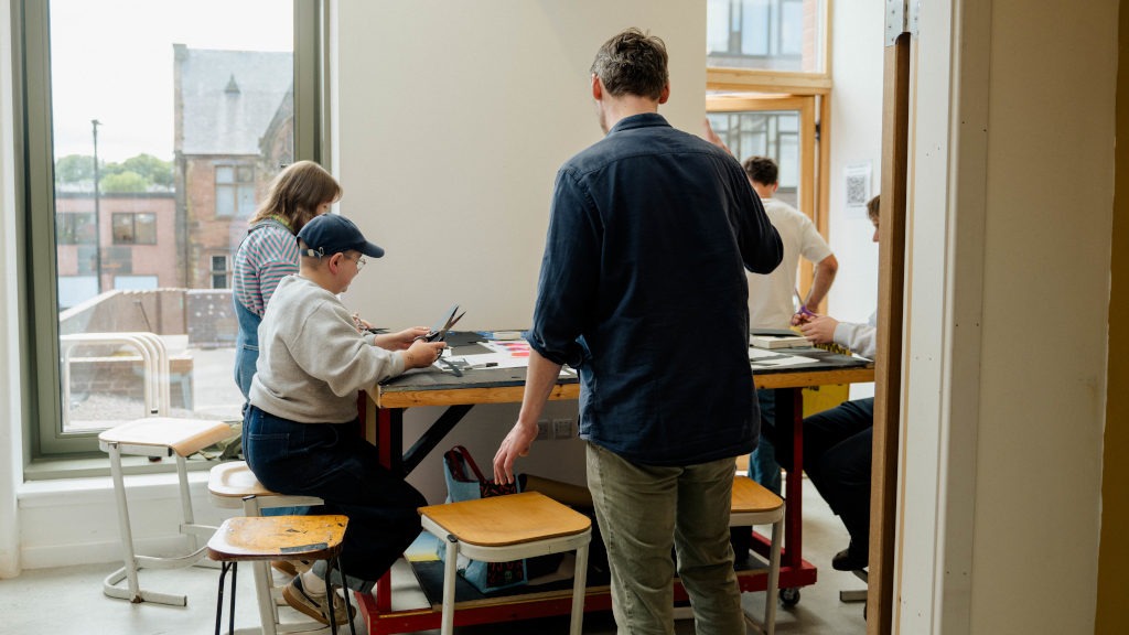Group of people gathered around a rectangular table in a bright room with large windows. Some are seated and working with paper and scissors, while one person stands nearby assisting or observing. The table holds various craft materials, and the scene conveys a collaborative workshop.