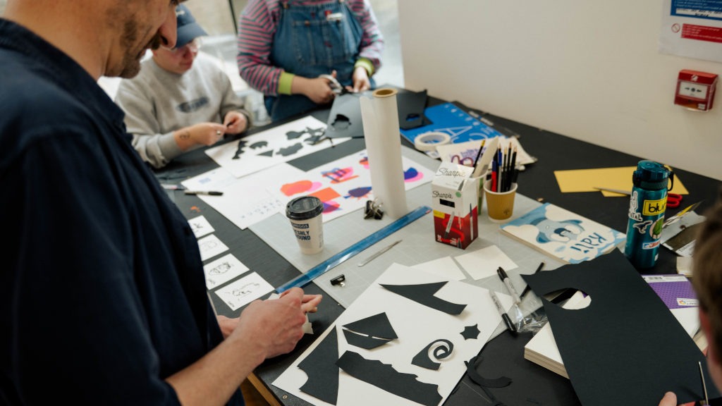 Several people seated at a table in a workshop or classroom, working on arts and crafts. The table is covered with black and white paper cutouts, scissors, markers, glue sticks, a roll of paper towels, and a coffee cup. One person cuts black paper while others assemble or draw on paper. Colourful paper designs and sketches are also visible, suggesting a collaborative collage or paper art project.
