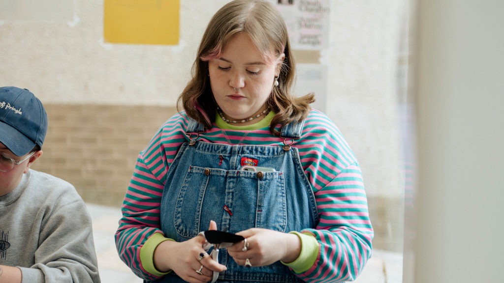 Person wearing a striped pink and green long‑sleeve shirt under blue denim overalls, with shoulder‑length hair featuring pink highlights. They are accessorised with rings and a choker necklace and appear focused on an object in their hands. Another person in a grey sweatshirt and blue cap is partially visible to the left. The background includes posters on the wall and a window, suggesting an indoor workshop.