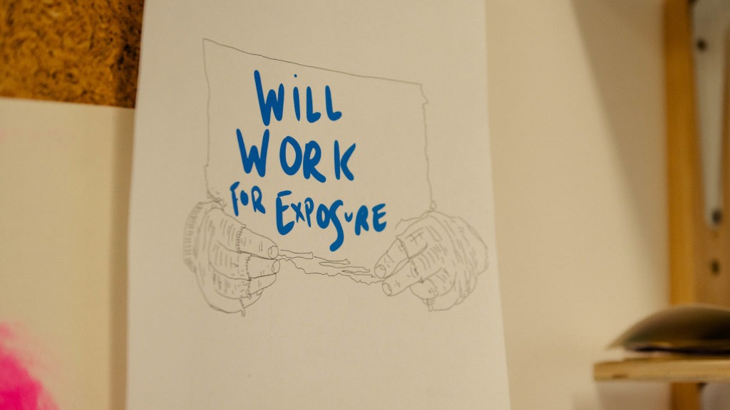 Hand‑drawn illustration pinned to a corkboard, showing two hands holding a sign with the words “WILL WORK FOR EXPOSURE” in bold blue letters. The drawing is in a sketch style with thin lines for the hands and sign, while the text is prominently coloured and stylised.